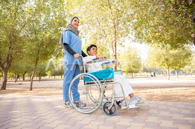 الاستمتاع بالأجواء الصيفيه في أوقات الفراغ، ممرضة عربية خليجيه سعودية ترتدي اللباس الخاص بالعمل برفقة ولد سعودي مقعد من ذوي الهمم يرتدي الثوب التقليدي يجلس على كرسي متحرك في إحدى الحدائق العامة، مفهوم الرعاية والعناية بالمرضى والمعاقين، التنزه في الهواء الطلق