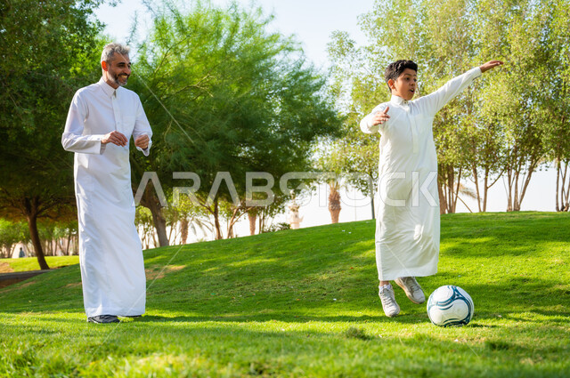إيماءات الحماس والنشاط، ممارسة رياضة كرة القدم, أب عربي سعودي خليجي يرتدي الزي التقليدي ويلعب مع ابنه في الحديقة العامة,  قضاء إجازة ممتعة في المنتزهات, صورة الجسم بالطول الكامل, الطبيعة الخضراء في المملكة العربية  السعودية.