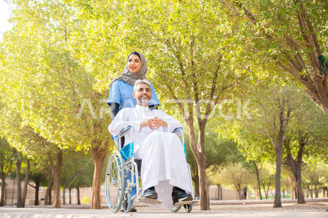 التحسين من نفسية المرضى  بالتجول في الهواء الطلق، قوة العزيمة والتحدي عند أصحاب الهمم, رجل كبير السن عربي خليجي سعودي مبتسم مقعد يرتدي الثوب التقليدي يجلس على كرسي متحرك برفقة ممرضته الخاصة في إحدى الحدائق العامة، شعور المعاقين بالإنتماء والتقدير في السعوديه