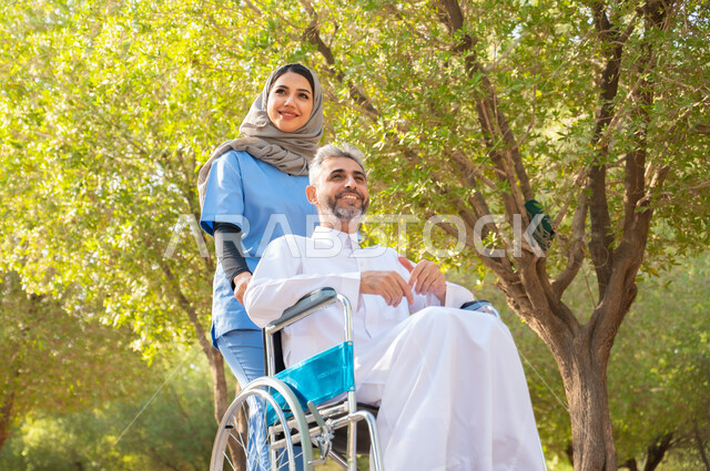 شعور المعاقين بالإنتماء والتقدير في السعوديه ,التحسين من نفسية المرضى بالتجول في الهواء الطلق، قوة العزيمة والتحدي عند أصحاب الهمم, رجل كبير السن عربي خليجي سعودي مبتسم مقعد يرتدي الثوب التقليدي يجلس على كرسي متحرك برفقة ممرضته الخاصة في إحدى الحدائق العامة