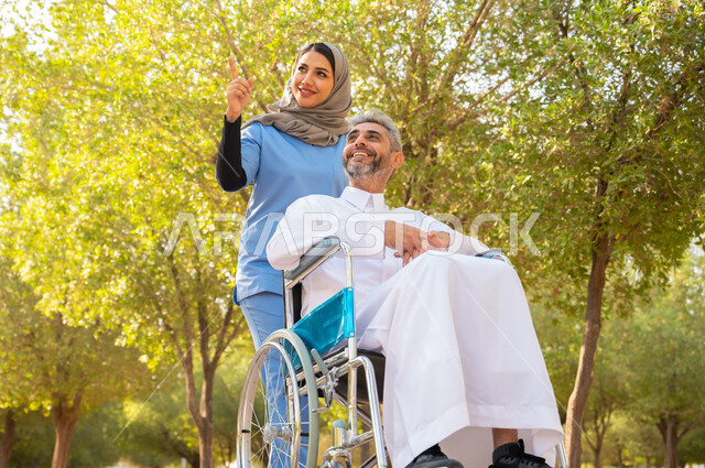 الاشارة إلى مكان ما ،شعور المعاقين بالتقدير و الإنتماء في السعوديه ،التحسين من نفسية المرضى بالتنزه في الهواء الطلق، قوة العزيمة والتحدي عند أصحاب الهمم, رجل مسن عربي خليجي سعودي مقعد يرتدي الثوب التقليدي يجلس على كرسي متحرك برفقة ممرضته الخاصة في أحد الحدائق