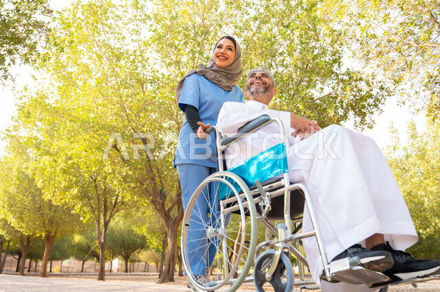 التسامر والضحك مع المرضى والتحسين من نفسيتهم، قوه الارادة و العزيمة عند أصحاب الهمم، شعور المعاقين بالإنتماء والتقدير والإحترام في السعوديه ،رجل كبير السن عربي خليجي سعودي مبتسم مقعد يرتدي الثوب التقليدي يجلس على كرسي متحرك برفقة ممرضته الخاصة في إحدى المنتزهات