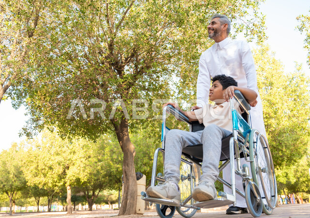 A family atmosphere full of love, weakness in motor and physical abilities, people of determination feeling comfortable and reassured in their environment, an elderly Saudi Arabian Gulf man wearing a traditional dress with his son in a park enjoying the outdoors, a disabled Saudi boy sitting in a wheelchair in a park
