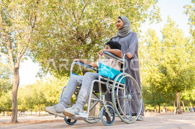 الضعف بالقدرات الحركية والجسدية، شعور ذوي الهمم بالراحه والطمأنينه في بيئتهم, أم عربيه خليجيه سعودية محجبة ترتدي العباءة الخليجيه برفقه ابنها في إحدى الحدائق للاستمتاع بالهواء الطلق، ولد سعودي مقعد يجلس على كرسي متحرك في أحد المنتزهات، اجواء عائلية مليئة بالحب