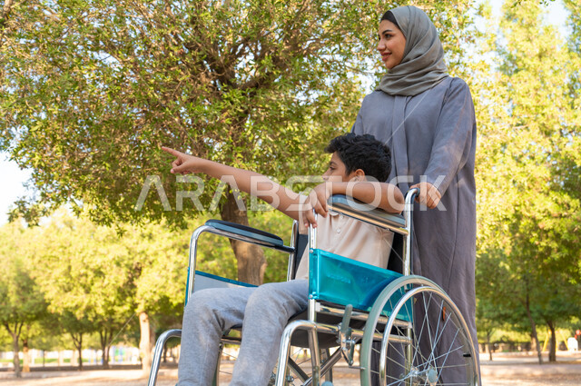 الحب والعطف بين أفراد الأسرة، مفهوم العنايه ورعاية المرضى والمعاقين، امرأة عربيه خليجيه سعودية محجبة ترتدي العباءة الخليجيه برفقه ابنها في إحدى المنتزهات، ولد سعودي مقعد يجلس على كرسي متحرك ويشير إلى شي ما، مساعدة مرضى ذوي الإعاقة الحركية على التنقل بسهولة