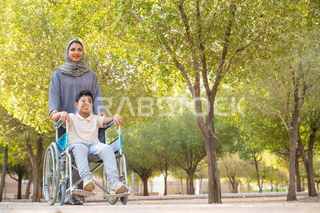 منح مرضى ذوي الإعاقة الحركية طاقة ايجابية، شعور ذوي الهمم بالراحه والطمأنينه في بيئتهم, امرأة عربيه خليجيه سعودية محجبة ترتدي العباءة الخليجيه برفقه ابنها في إحدى الحدائق للاستمتاع بالهواء الطلق، ولد سعودي مقعد يجلس على كرسي متحرك في أحد المنتزهات