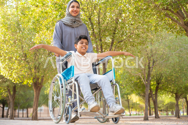 معدات تساعد مرضى ذوي الإعاقة الحركية على التنقل بسهولة, شعور ذوي الهمم بالراحه والطمأنينه في بيئتهم, أم عربيه خليجيه سعودية محجبة ترتدي العباءة الخليجيه برفقه ابنها في إحدى المنتزهات للاستمتاع بالهواء الطلق، ولد سعودي مقعد يجلس على كرسي متحرك يمد يديه جانبا