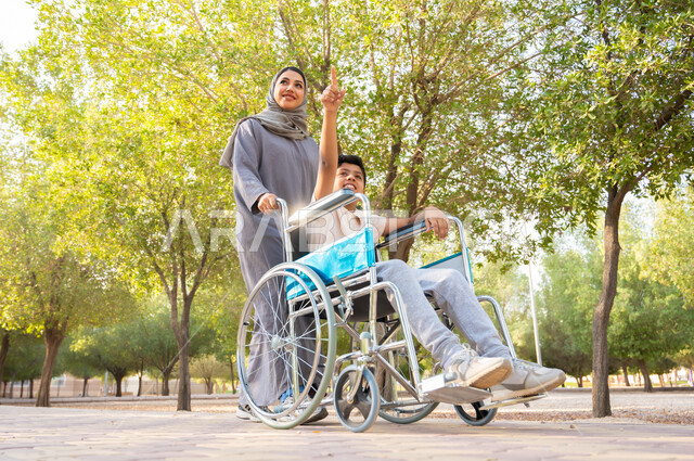 مساعدة مرضى ذوي الإعاقة الحركية على التنقل بسهولة, شعور ذوي الهمم بالراحه والطمأنينه في بيئتهم, امرأة عربيه خليجيه سعودية محجبة ترتدي العباءة الخليجيه برفقة ابنها في إحدى الحدائق، ولد سعودي مقعد يجلس على كرسي متحرك ويشير إلى شي ما، العطف والحب بين أفراد الأسرة