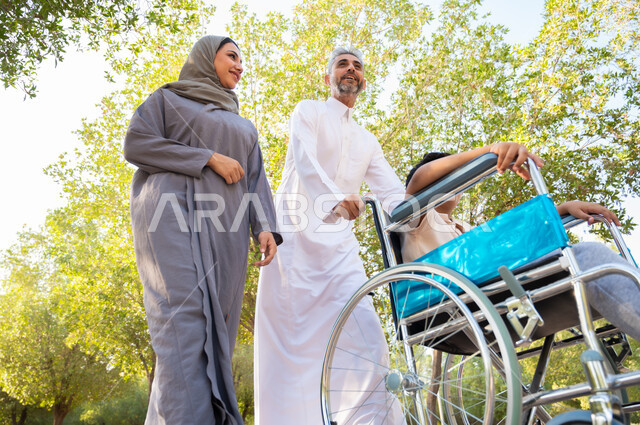 الاعتناء بالأبناء المعاقين والتحسين من نفسيتهم، عائلة عربيه خليجيه سعودية تقوم بالتنزه في إحدى الحدائق العامة ،ولد سعودي من ذوي الهمم يجلس على كرسي متحرك برفقة والديه في الخارج للاستمتاع بالعطلة الصيفيه، أجواء عائلية مليئة بالحب، الحنان والعطف بين أفراد الأسرة