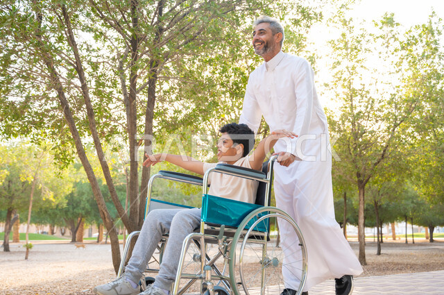 الاهتمام والرعاية بالأولاد ،أدوات تساعد مرضى ذوي الإعاقة الحركية على التنقل بسهولة, تحسين حياة أصحاب الهمم وتأمين احتياجاتهم الخاصة, رجل مسن عربي خليجي سعودي يرتدي الزي التقليدي برفقة ابنه في إحدى الحدائق، ولد سعودي مقعد يجلس على كرسي متحرك يمد يديه جانبا