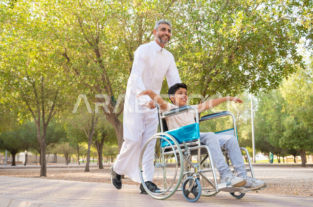 تحسين حياة أصحاب الهمم وتأمين احتياجاتهم الخاصة, معدات تساعد مرضى ذوي الإعاقة الحركية على التنقل بسهولة, رجل مسن عربي خليجي سعودي يرتدي الزي التقليدي برفقة ابنه في إحدى الحدائق للاستمتاع بالهواء الطلق، ولد سعودي مقعد يجلس على كرسي متحرك يمد يديه جانبا