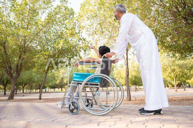 تأمين الاحتياجات الخاصة لأصحاب الهمم, الاهتمام والرعاية بالأولاد ،شعور ذوي الاعاقة بالطمأنينه في بيئتهم, صورة من الخلف لرجل مسن عربي خليجي سعودي يرتدي الثوب التقليدي برفقة ابنه في إحدى الحدائق، ولد سعودي مقعد يجلس على كرسي متحرك في أحد المنتزهات يشير إلى شيء ما