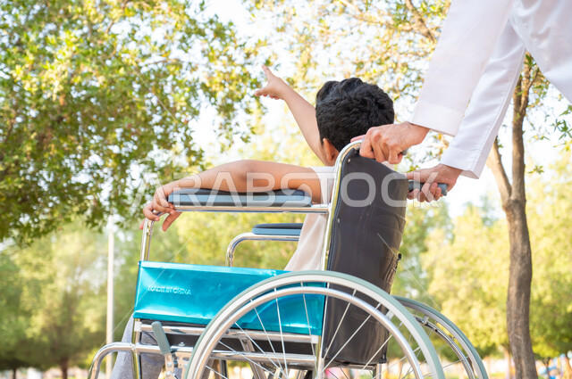 شعور ذوي الهمم بالراحه والطمأنينه في بيئتهم, التحسين من نفسية الأبناء ومنحهم الطاقة الايجابية، صورة من الخلف لرجل مسن عربي خليجي سعودي يرتدي الثوب التقليدي برفقة ابنه في إحدى الحدائق، ولد سعودي مقعد يجلس على كرسي متحرك في أحد المنتزهات يشير إلى شيء ما