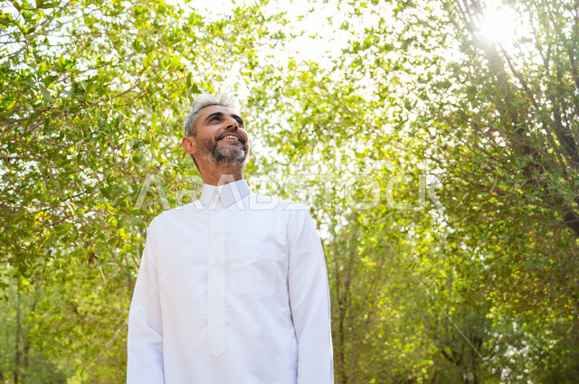 الاسترخاء والراحة في الإجازة، إيماءات التأمل والنظر إلى شيء ما، رجل كبير السن عربي سعودي خليجي مبتسم يرتدي الثوب التقليدي ويتنزه في الهواء الطلق، الاستجمام والاستمتاع بالمناظر الطبيعية، قضاء عطلة صيفية ممتعة, الطبيعة الخضراء في المملكة العربية السعودية.