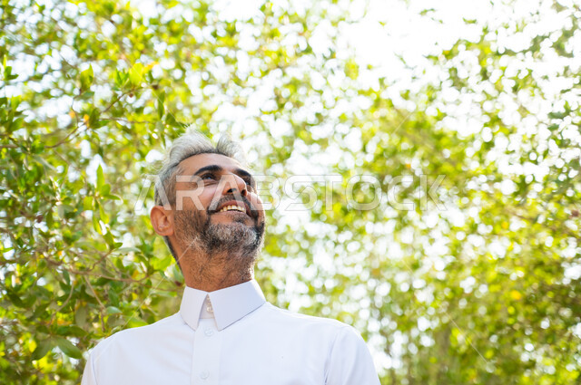 الاستجمام والاستمتاع بالمناظر الطبيعية، إيماءات التأمل والنظر إلى شيء ما، رجل كبير السن عربي سعودي خليجي مبتسم يرتدي الثوب التقليدي ويتنزه في الهواء الطلق، الاسترخاء والراحة في الإجازة، قضاء عطلة صيفية ممتعة, الطبيعة الخضراء في المملكة العربية السعودية.