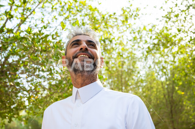 الشعور بالراحة والاسترخاء، رجل شايب عربي سعودي خليجي مبتسم يرتدي الثوب التقليدي وينظر إلى شيء ما، الاستجمام والتنزه في الهواء الطلق، قضاء أوقات ممتعة في العطلة الصيفية، الطبيعة الخضراء في المملكة العربية السعودية.