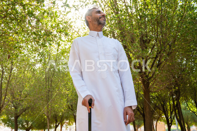 نزهة في الطبيعة و الهواء الطلق، الخروج إلى أحد الحدائق العامة للترفيه عن النفس، الاستمتاع بالأجواء الصيفيه في إحدى المنتزهات، رجل كبير السن عربي خليجي سعودي يرتدي الثوب التقليدي يرتكز على عكاز لسهولة التنقل بإيماءات السعادة، معدات تساعد على المشي.