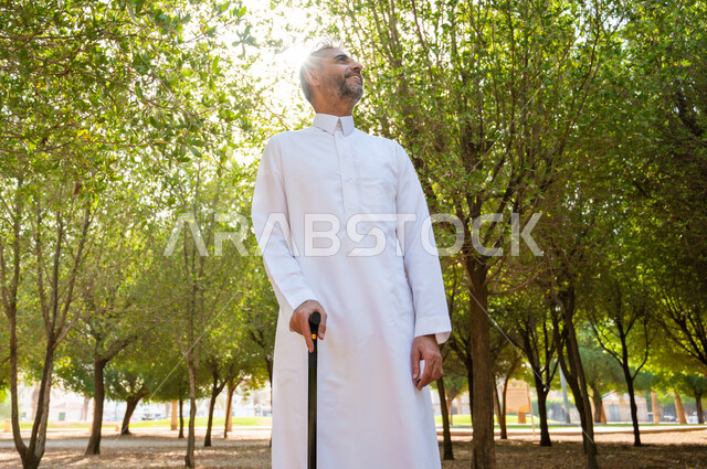 الاستجمام بالطبيعة واستنشاق الهواء الطلق، الخروج إلى أحد الحدائق العامة للترفيه عن النفس، الاستمتاع بالأجواء الصيفيه في إحدى المنتزهات، رجل شايب عربي خليجي سعودي يرتدي الثوب التقليدي يرتكز على عكاز لسهولة التنقل بإيماءات السعادة، معدات تساعد على المشي.