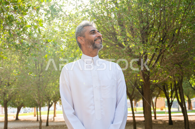 Hiking and walking in public parks to breathe the fresh air, a smiling old Saudi Arabian Gulf man wearing a traditional dress and looking at something, relaxing, contemplating and feeling comfortable, spending enjoyable times in the summer vacation, the green nature in the Kingdom of Saudi Arabia