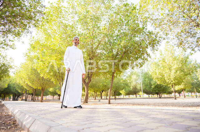 Going to one of the public parks to entertain oneself, taking a walk in nature and the open air, enjoying the summer atmosphere in one of the parks, an elderly Saudi Arabian Gulf man wearing a traditional dress, resting on a crutch for easy movement with happy gestures, equipment that helps with walking