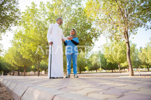 التحادث والتسامر بإيماءات الفرح، مفهوم الخدمات الطبية والعناية الصحية، ممرضة عربية خليجية سعودية تمسك يد مريضها وتساعده على المشي، رجل كبير السن يرتدي الزي التقليدي يستند على العكاز يتنزه في الحديقة، صورة الجسم بالطول الكامل، الطبيعة الخضراء في المملكة