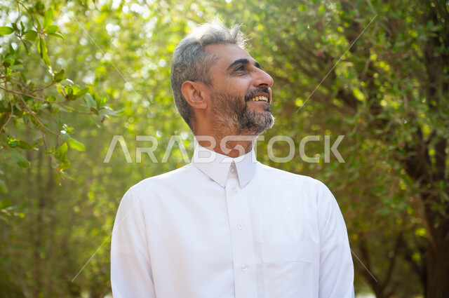 الاستجمام والتأمل والشعور بالراحة، رجل كبير السن عربي سعودي خليجي مبتسم يرتدي الثوب التقليدي وينظر إلى شيء ما، قضاء أوقات ممتعة في العطلة الصيفية، التنزه و المشي في الحدائق العامة لاستنشاق الهواء الطلق، الطبيعة الخضراء في المملكة العربية السعودية.