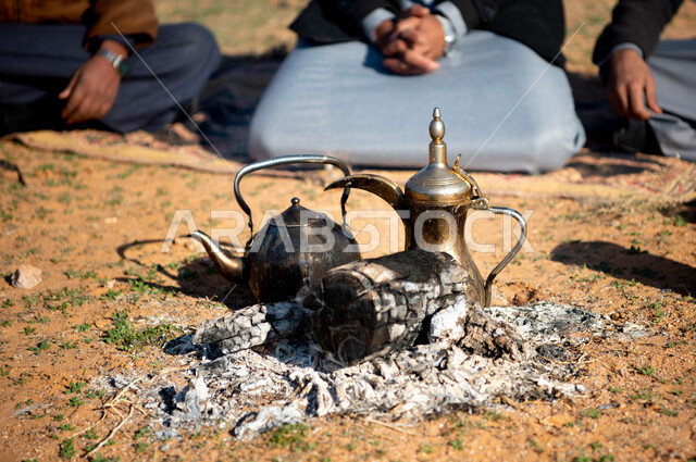 الاستمتاع بشرب القهوة العربية, صورة مقربة لدلة قهوة وابريق شاي على موقد حطب في الصحراء السعودية، الجلسات السعودية التقليدية الشعبية، قضاء أوقات ممتعة في الأجواء الصحراوية, رجال عرب خليجيين سعوديين في رحلة تخييم برية، كشتة بر شبابية في وقت النهار.