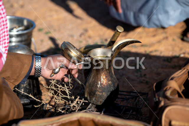 الجلسات السعودية التقليدية الشعبية، الاستمتاع بشرب القهوة العربية, صورة مقربة ليد رجل عربي سعودي خليجي يمسك دلة قهوة ويضعها على موقد حطب في الصحراء، قضاء أوقات ممتعة في الأجواء الصحراوية, رحلة تخييم برية وكشتة بر شبابية في وقت النهار.