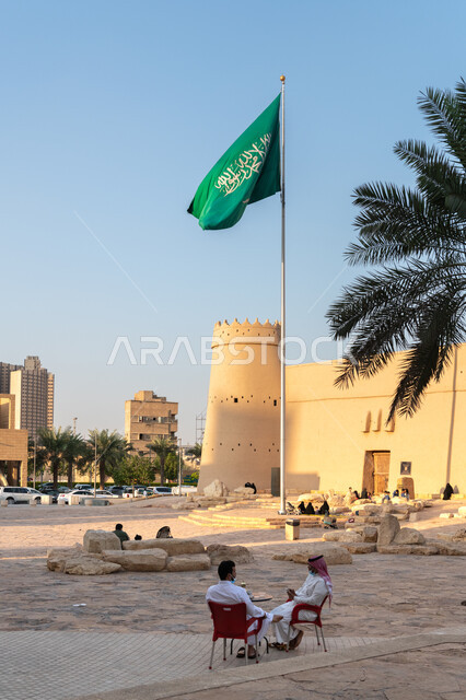 رفع علم السعودية في ساحات منطقة قصر الحكم في اليوم الوطني السعودي23 سبتمبر في مدينة الرياض، الملتقى الاجتماعي لسكان العاصمة، المعالم والمواقع الأثرية السياحية، المباني الطينية والعمارات القديمة، صورة من الخلف لرجلان خليجيان سعوديان يجلسان في ساحة متحف المصمك