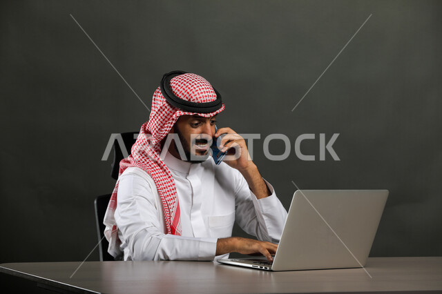 إيماءات الفرح بتحقيق أهداف الشركه، بورتريه لرجل أعمال عربي  سعودي خليجي مبتسم يرتدي الشماغ والثوب التقليدي يجري مكالمة هاتفيه في مقر العمل، مهن وأعمال تجارية عن بعد، استخدام الهاتف المحمول، مراجعة سير المخططات عبر اللابتوب، خلفية رمادية .