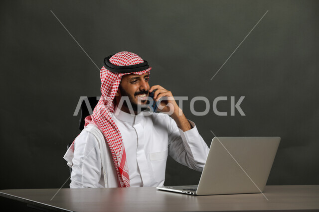  مهن وأعمال تجارية عن بعد، إيماءات الفرح بتحقيق أهداف الشركه، بورتريه لرجل أعمال عربي  سعودي خليجي مبتسم يرتدي الشماغ والثوب التقليدي يجري مكالمة هاتفيه في مقر العمل، استخدام الهاتف المحمول، عقد صفقات واتفاقيات عبر الإنترنت، خلفية رمادية .