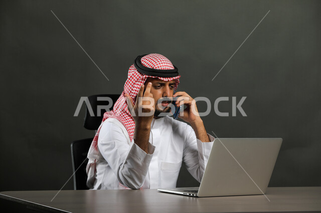 Arguments and discussions about projects using a mobile phone, gestures of annoyance and dissatisfaction, a portrait of a Saudi-Gulf Arab businessman wearing a shemagh and traditional dress making a phone call at the workplace, making deals and agreements over the Internet, a gray background