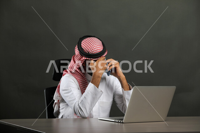 إيماءات التفكير والتأمل لوضع الخطط والاستراتيجيات، بورتريه لرجل أعمال عربي  سعودي خليجي يرتدي الشماغ والثوب التقليدي يجري مكالمة هاتفيه في مقر العمل، تشاور ومحادثات باستخدام الهاتف المحمول، عقد صفقات واتفاقيات عبر الإنترنت، خلفية رمادية .