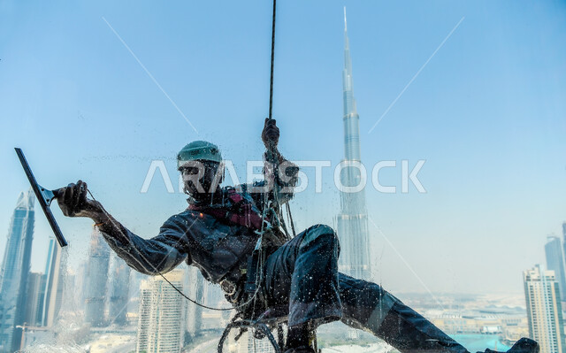 مهن ووظائف شاقة وخطيره ,الادوات والمعدات اللازمة لإزالة الغبار عن الزجاج, رجل عربي خليجي سعودي يرتدي اللباس الخاص في التسلق لتنظيف الواجهات الزجاجية لأحد الابراج في مدينة دبي, خلفية المباني وناطحات السحاب في الامارات العربيه المتحدة