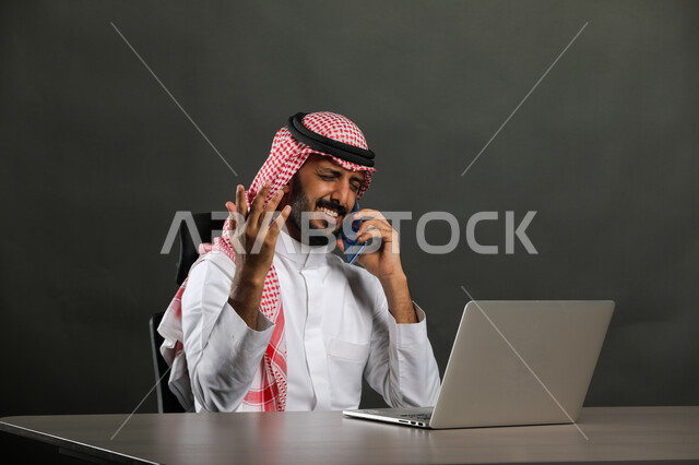 إيماءات الملل والتعب من تكرار الأخطاء في العمل،مراجعة المخططات عبر جهاز الحاسوب، بورتريه لرجل أعمال عربي  سعودي خليجي يرتدي الشماغ والثوب التقليدي يجري مكالمة هاتفيه باستخدام الهاتف المحمول، الخسارة والفشل في تحقيق الأهداف، خلفية رمادية .