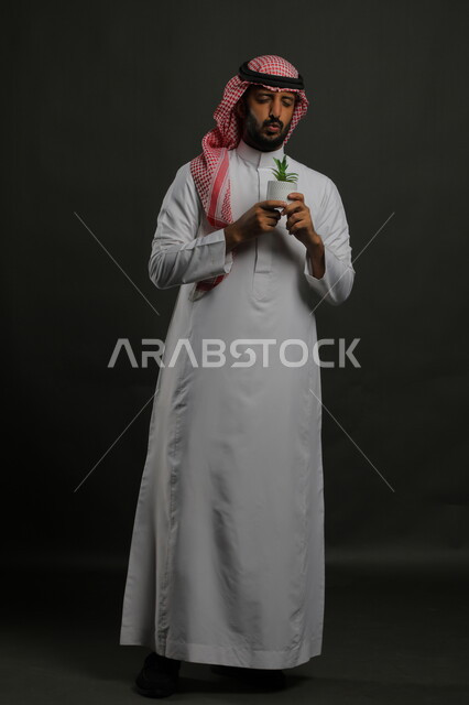 العناية بنباتات الزينة, الاهتمام بالشتلات المنزلية, بورتريه لرجل عربي خليجي سعودي يرتدي الثوب التقليدي والشماغ يحمل بيده حوض زراعي صغير, الوقوف باستقامة بإيماءات الإعجاب بالنبات, صورة الجسم بالطول الكامل, خلفية سوداء