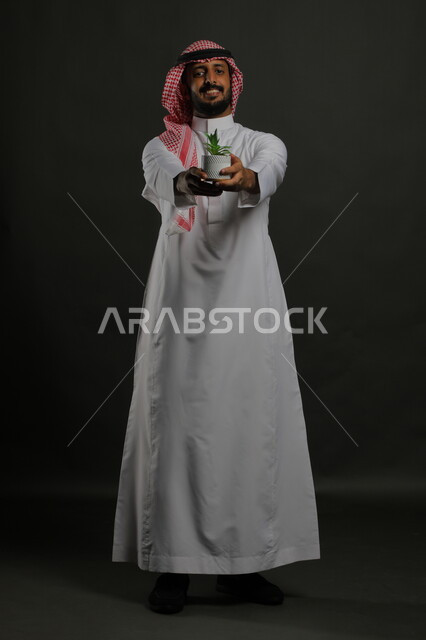 Taking care of home seedlings, taking care of ornamental plants, a portrait of a smiling Saudi Arabian Gulf man wearing a traditional thobe and shemagh, holding a small agricultural basin in his hand, standing up straight and looking at the camera with gestures of pleasure and joy, full-length body portrait, black background