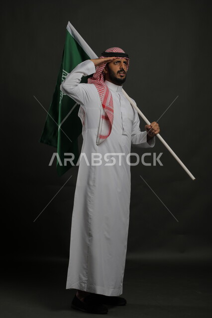 إيماءات الجدية والحزم، الاحتفال باليوم الوطني السعودي 23 سبتمبر، الوقوف بانضباط وإلقاء التحية للوطن والعلم، إحياء ذكرى يوم العلم  11 مارس، بورتريه لشاب عربي خليجي سعودي يرتدي الشماغ والثوب التقليدي يحمل بيده علم المملكة، صورة الجسم بالطول الكامل , خلفية رمادية