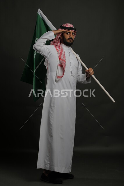  الاحتفال باليوم الوطني السعودي 23 سبتمبر، الوقوف بانضباط وإلقاء التحية للوطن والعلم، إحياء ذكرى يوم العلم  11 مارس، بورتريه لشاب عربي خليجي سعودي يرتدي الشماغ والثوب التقليدي ينظر إلى الكاميرا يحمل بيده علم المملكة، صورة الجسم بالطول الكامل , خلفية رمادية