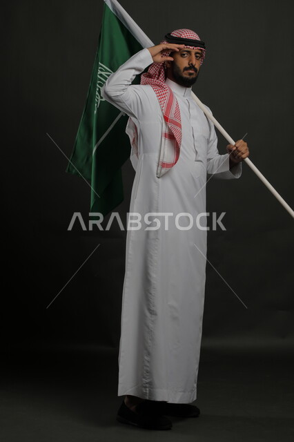 الوقوف بانضباط وإلقاء التحية للوطن والعلم، إحياء ذكرى يوم العلم  11 مارس، الاحتفال باليوم الوطني السعودي 23 سبتمبر، بورتريه لشاب عربي خليجي سعودي يرتدي الشماغ والثوب التقليدي ينظر إلى الكاميرا يحمل بيده علم المملكة، صورة الجسم بالطول الكامل , خلفية رمادية