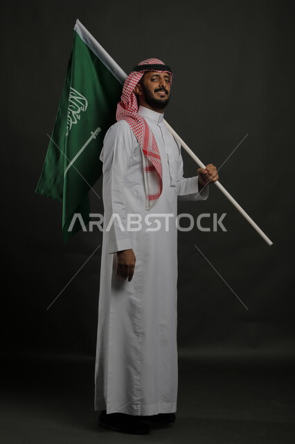 إيماءات الفرح والصمود في يوم العلم  11 مارس، الوقوف باستقامة والنظر إلى الكاميرا، السعادة في الاحتفال باليوم الوطني السعودي 23 سبتمبر، بورتريه لشاب عربي خليجي سعودي مبتسم يرتدي الشماغ والثوب التقليدي يحمل بيده علم المملكة، صورة الجسم بالطول الكامل , خلفية رمادية