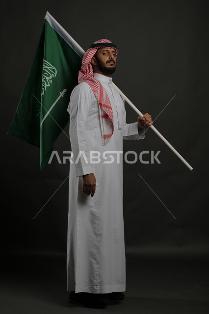 الصمود أثناء الاحتفال باليوم الوطني السعودي 23 سبتمبر، بورتريه لرجل عربي خليجي سعودي يرتدي الشماغ والثوب التقليدي يحمل بيده علم المملكة ، الوقوف باستقامة والنظر إلى الكاميرا بإيماءات الثقة بالنفس في يوم العلم  11 مارس ، صورة الجسم بالطول الكامل , خلفية رمادية