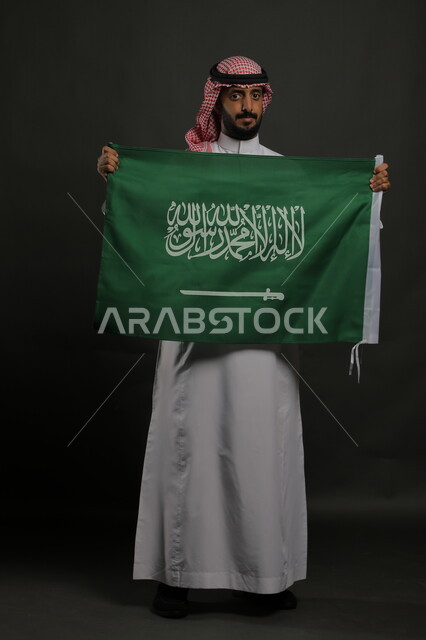 الجد والحزم في مسك راية التوحيد والإسلام، الاعتزاز بذكرى اليوم الوطني السعودي 23 سبتمبر، بورتريه لشاب عربي خليجي سعودي يرتدي الشماغ والثوب التقليدي يحمل بيده علم المملكة ، الاحتفال بيوم العلم  11 مارس ، صورة الجسم بالطول الكامل , خلفية رمادية