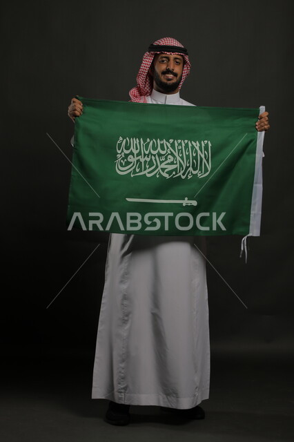 الفرح والاعتزاز بذكرى اليوم الوطني السعودي 23 سبتمبر، راية التوحيد والإسلام، بورتريه لرجل عربي خليجي سعودي مبتسم يرتدي الشماغ والثوب التقليدي يحمل بيده علم المملكة ، الاحتفال بيوم العلم  11 مارس ، صورة الجسم بالطول الكامل , خلفية رمادية