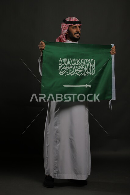 راية التوحيد والإسلام، الاعتزاز بذكرى اليوم الوطني السعودي 23 سبتمبر، بورتريه لرجل عربي خليجي سعودي مبتسم يرتدي الشماغ والثوب التقليدي يحمل بيده علم المملكة ، الاحتفال بيوم العلم  11 مارس ، صورة الجسم بالطول الكامل , خلفية رمادية