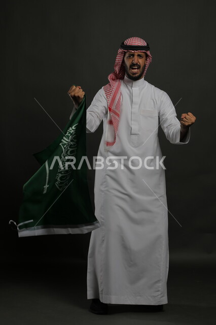 التشجيع والحماس أثناء الاحتفال باليوم الوطني السعودي 23 سبتمبر، بورتريه لشاب عربي خليجي سعودي يرتدي الشماغ والثوب التقليدي يحمل بيده علم المملكة ، النظر إلى الكاميرا بإيماءات الانتصار والفوز في يوم العلم  11 مارس ، صورة الجسم بالطول الكامل , خلفية رمادية