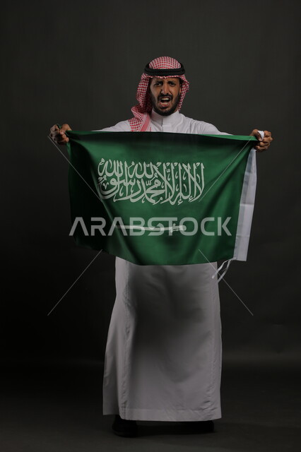 مشاعر الحماس أثناء الاحتفال باليوم الوطني السعودي 23 سبتمبر، بورتريه لرجل عربي خليجي سعودي يرتدي الشماغ والثوب التقليدي يحمل بيده علم المملكة ، النظر إلى الكاميرا بإيماءات الفرح والانتصار في يوم العلم  11 مارس ، صورة الجسم بالطول الكامل , خلفية رمادية