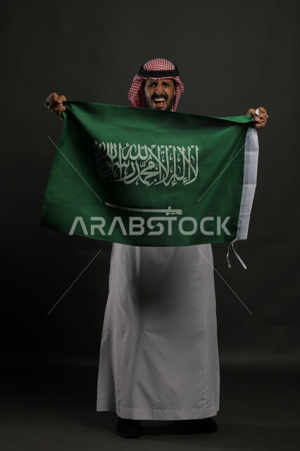 إيماءات التشجيع والحماس في الاحتفال باليوم الوطني السعودي 23 سبتمبر، النظر إلى الكاميرا بإيماءات الفوز والنصر في يوم العلم  11 مارس، بورتريه لشاب عربي خليجي سعودي يرتدي الشماغ والثوب التقليدي يحمل بيده علم المملكة، صورة الجسم بالطول الكامل, خلفية رمادية