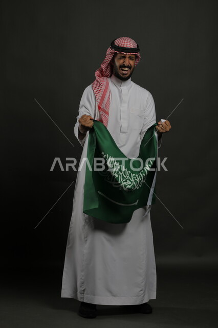 الشعور بالسعادة والفرح في الاحتفال باليوم الوطني السعودي 23 سبتمبر، بورتريه لرجل عربي خليجي سعودي يرتدي الشماغ والثوب التقليدي يحمل بيده علم المملكة ،إيماءات الفوز والنصر في يوم العلم  11 مارس ، صورة الجسم بالطول الكامل , خلفية رمادية