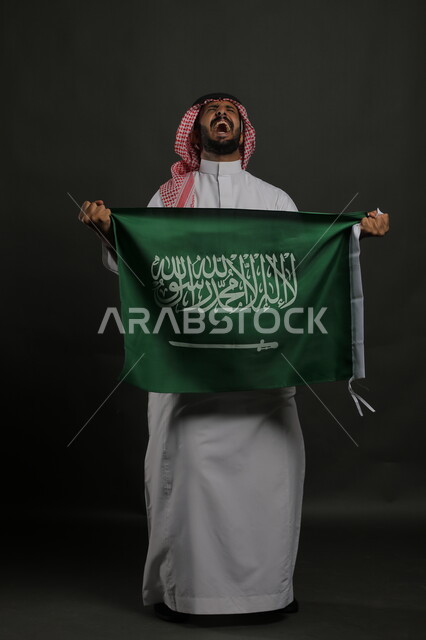 الشكر والامتنان لله على النصر في اليوم الوطني السعودي 23 سبتمبر، بورتريه لرجل عربي خليجي سعودي يرتدي الشماغ والثوب التقليدي يحمل بيده علم المملكة ، إيماءات الالفرح والسرور في يوم العلم  11 مارس ، صورة الجسم بالطول الكامل , خلفية رمادية